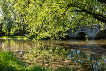 Sunlit river scene with an arched stone bridge green trees and reflections on the water creating a...