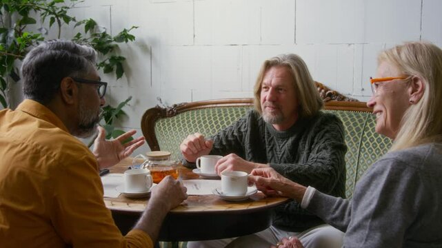 Medium close-up of mature Asian man in telling funny life story or anecdote to Caucasian couple of friends, who are listening and laughing heartily, while enjoying lunchtime meeting in cozy cafe