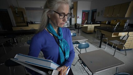 Closeup of mature woman teacher, dressed professionally, stands in an empty school biology laboratory, holding books and looking sad and unhappy as she prepares for her upcoming lesson.
