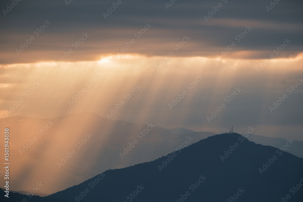 Poster sunrays break through clouds over hazy mountains silhouetting peaks and adding depth to the landscap