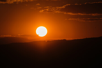 Fiery Sunset: The sun descends over shadowy hills painting the sky with vibrant orange hues as...