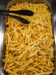 Metal tray is filled with a large quantity of golden french fries, ready to be served with black tongs resting on top