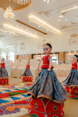 Elaborate traditional dolls adorn event space. Dressed in colorful garments, they rest on red ornamented bases, adding cultural charm to the setting.