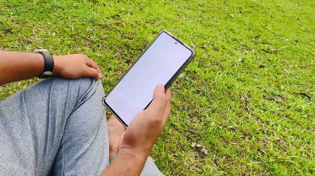 Man Using Smartphone While Sitting on Green Grass in Park, Typing and Reading Messages

