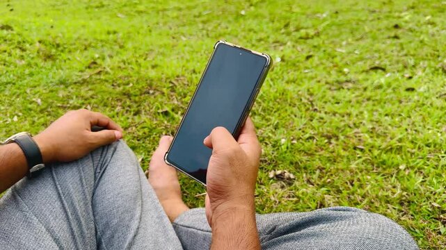 Man Using Smartphone While Sitting on Green Grass in Park, Typing and Reading Messages

