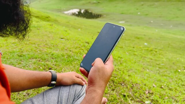 Man Using Smartphone While Sitting on Green Grass in Park, Typing and Reading Messages

