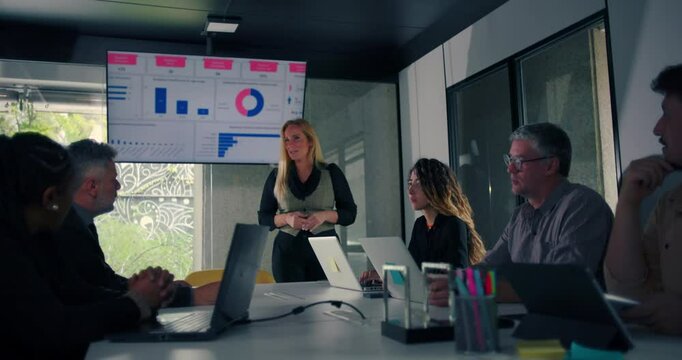 Blonde businesswoman delivering presentation to engaged colleagues seated around conference table with data screen in modern office