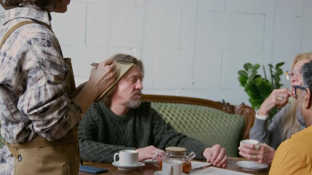 Medium close-up of three diverse middle-aged companions looking at menu while socializing together in cafe, drinking tea, asking waitress for dessert recommendations