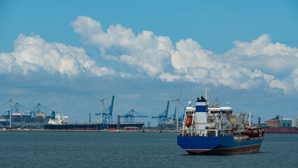 Blue cargo ship in industrial harbor with cranes, wind turbines, and cloudy sky