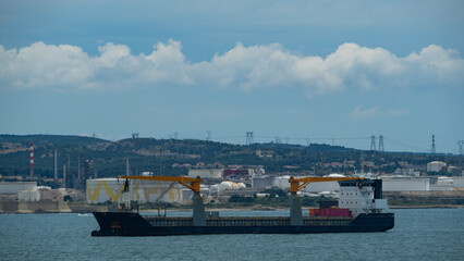 Bulk carrier vessel sailing on the sea with industrial facilities and hills in the background under...