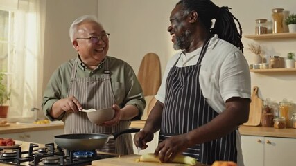 Two diverse male smiling senior friends wearing aprons cooking together in modern kitchen, enjoying their time and sharing a laugh while preparing a meal - Powered by Adobe