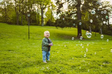 Little 2 year old boy running on green grass with soap bubbles. Toddler boy enjoying a walk in nature. Child having fun in city park.