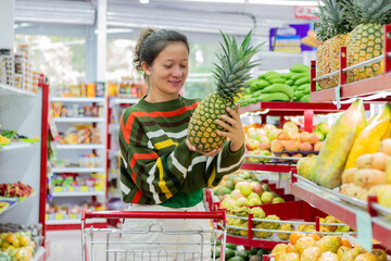 Smiling woman pushing a shopping cart and choosing a pineapple in the fruit and vegetable aisle of a grocery store, examining the quality of the produce before making a purchase