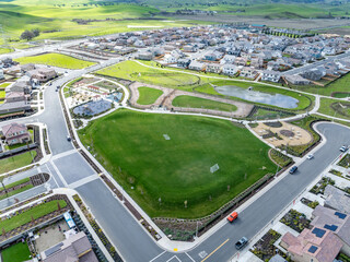 Aerial view of a suburban recreational area with soccer fields, walking paths, playground, pond, and surrounding homes set against rolling green hills on a clear day.