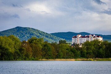 Panoramic view of modern residential buildings nestled at the base of lush green mountains, with a serene lake in the foreground. A perfect balance of urban living and natural beauty, ideal for concep