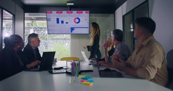 Businesswoman explains data chart to engaged team during office presentation while group of professionals seated around conference table watch screen