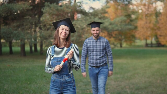 Two graduates rejoice after the ceremony, with one holding a diploma and the other walking behind in a park adorned with autumn colors.