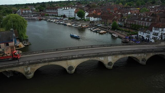 Aerial drone footage flying backwards over Henley Bridge, revealing the charming riverside town, boats moored along the banks of the River Thames, and vehicles, including a red truck, crossing the bri