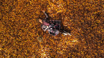 Aerial view  of an attractive young woman laying on the forest floor by natural sunlight surrounded by leaves in autumn fall colours. Elevated photo of woman is wearing coat and skirt .