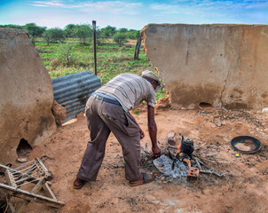 village african man cooking , tea and coffee pot on open fire in the yard at sunset, waiting for the food water  to cook boil