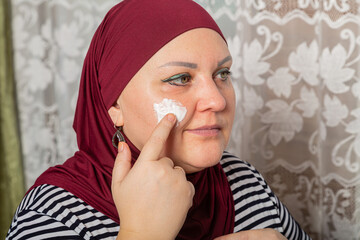 Muslim woman in red hijab sits in room near mirror and applies cream on face close-up