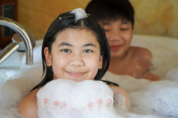 Photo of a smiling and happy Asian girl and brother playing with white foam in a bathtub at home.