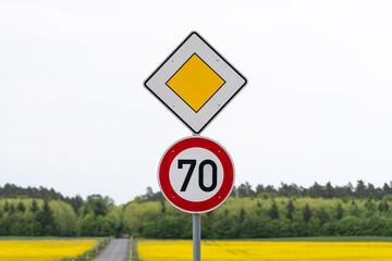 Main road traffic sign and 70 kilometers per hour speed limit next to a country road in Germany. Rural road in between agricultural fields in Europe.