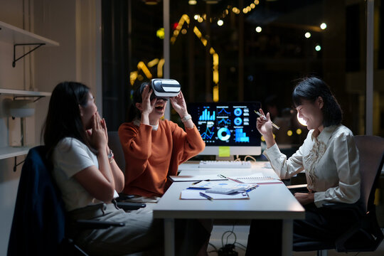 Asian businesswomen experiencing virtual reality headset in office meeting, exploring metaverse, presenting charts and graphs - Powered by Adobe