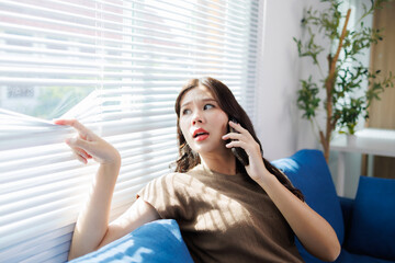Concerned young woman sitting on a blue couch, holding her phone to her ear, peeks through the...