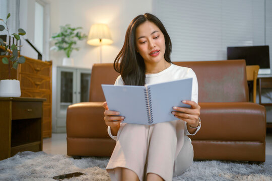 Smiling asian woman enjoying reading a book while sitting on the floor in her living room, with a couch in the background