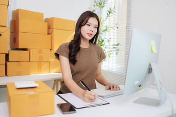 Young beautiful asian woman entrepreneur working from home checking orders and preparing packages for delivery, taking notes and using computer, surrounded by cardboard boxes