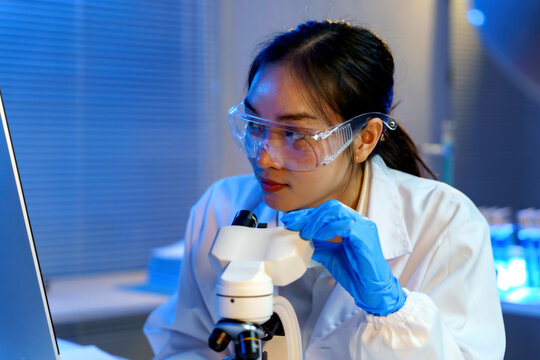 Young scientist wearing lab coat and protective glasses, adjusting microscope, performing scientific research in modern laboratory