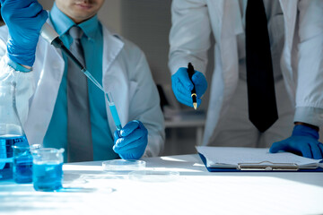 Two scientists wearing lab coats and gloves are conducting a chemical experiment with pipette and...