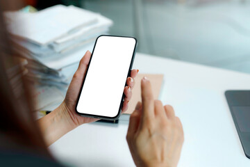 Businesswoman holding a smartphone with a blank white screen while working at an office desk, showcasing a modern workspace setup