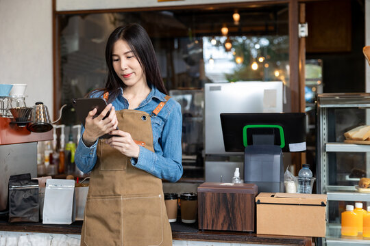 Young Asian female barista in brown apron using digital tablet behind coffee counter at café. Wearing denim shirt, standing near point of sale system and takeaway coffee cups, working attentively.