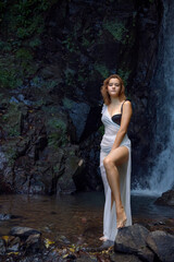 A girl in a transparent long white tunic with a bare shoulder stands against the backdrop of a waterfall in a grotto, putting one slender bare leg on a stone.