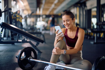 Happy athletic woman texting on smart phone while taking break during strength training in gym.