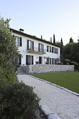 A white two-story house with a gravel pathway leading up to it, surrounded by lush greenery