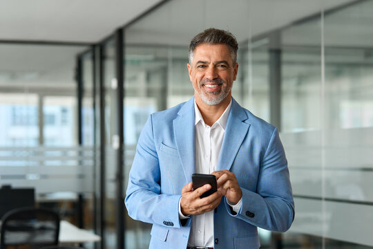 Happy mature 50 years old business man investor wearing suit standing in office using mobile phone. Smiling professional businessman of mid age holding smartphone looking at camera at work. Portrait. - Powered by Adobe