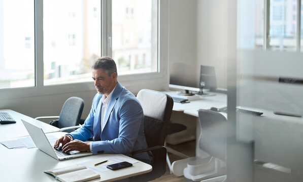 Busy older middle aged professional business man, senior executive manager wearing suit working on financial analysis using laptop computer in office. View through glass. Candid authentic photo.