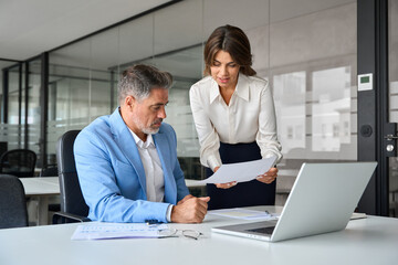 Obraz premium Two busy professionals man and woman business leaders of middle age checking accounting documents financial report talking working together on laptop computer in office at corporate meeting.