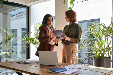 Two busy business women executive team talking using tab working together standing in office....