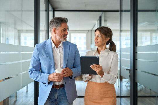Two busy business people executive team talking walking in office. Mature professional business man and woman partners working together having conversation in office using digital tab. Authentic photo