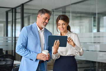 Two busy mature professional executives team business people talking using tablet computer having conversation standing in office, corporate managers working together with tab device.
