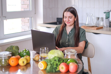 woman working on laptop in kitchen with vegetables and fruits on table healthy eating food blogging...