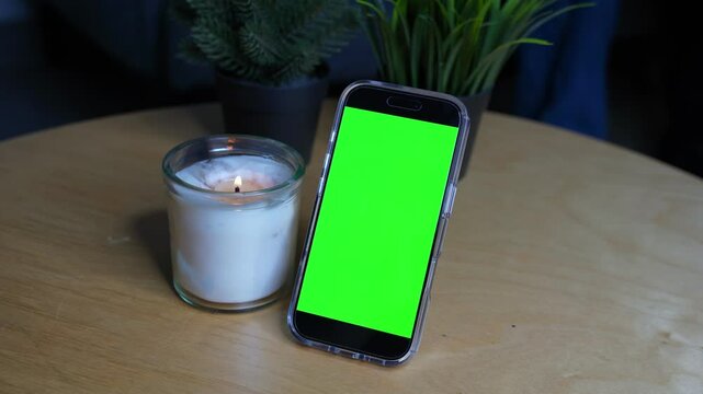4K footage of a smartphone with green screen beside a lit candle on a wooden table. Perfect for mockups, UI previews, or tech ads. Warm and modern indoor scene. Shot in Kuwait.