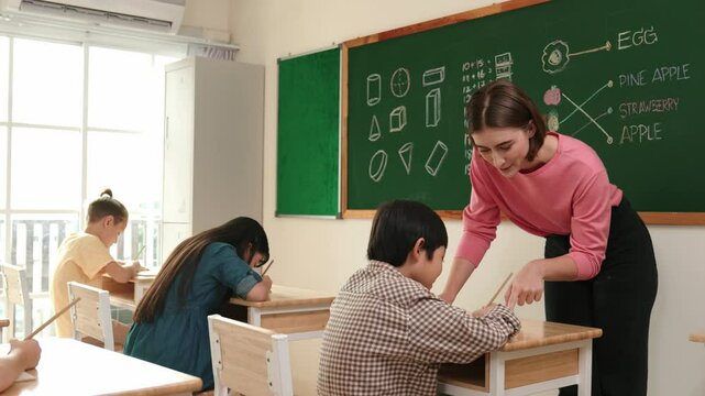 Smart teacher checking cute student homework while explaining idea at class. Attractive instructor walking around classroom while looking at notebook and talking about Math theory to child. Pedagogy.