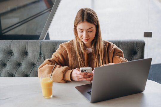 Young woman using smartphone and laptop in cafe, drinking orange juice - Powered by Adobe