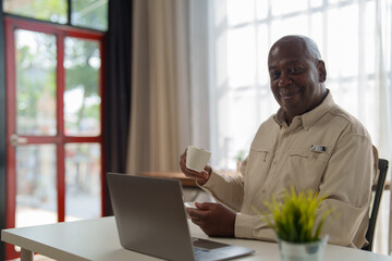 Happy african american businessman drinking coffee and using laptop while working from home office, smiling mature man enjoying break time with cup of coffee and using computer