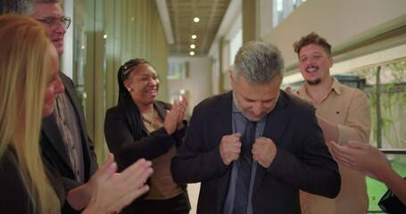 Grateful male professional smiling while receiving applause from supportive colleagues in office hallway, emotional moment of recognition and appreciation